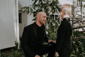 Two men in all black formal attire having a quiet moment among garden foliage.