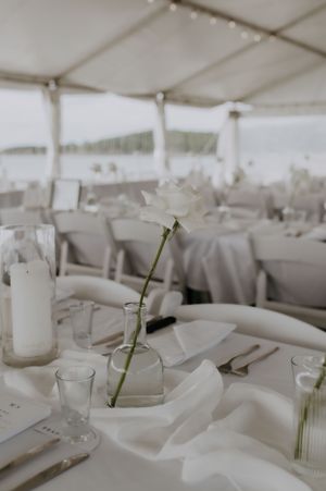 Elegant white wedding reception table setting under a tent with a minimalist single flower stem centerpiece.