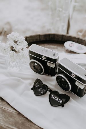Vintage-style cameras and heart-shaped sunglasses arranged on white fabric with small white flowers.