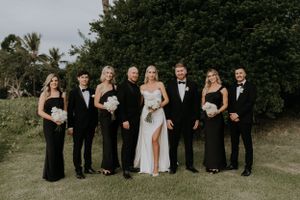 Wedding party in formal black attire poses outdoors against lush greenery at tropical venue.
