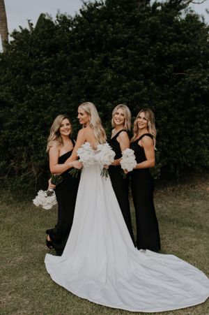 Bridesmaids in elegant black dresses gather around bride wearing flowing white gown with long train.