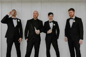 Four groomsmen in formal black tuxedos pose casually against a white wall.