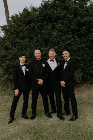 Groomsmen in black tuxedos pose together against green foliage backdrop.