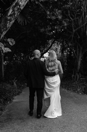A romantic black and white wedding photo of a couple walking down a garden path surrounded by lush foliage.