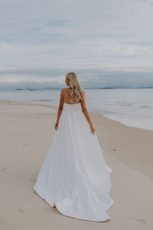 A bride in a flowing white wedding dress stands on a serene beach looking out at the ocean.