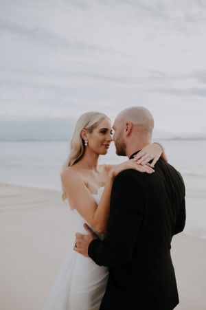Romantic beach wedding portrait of a couple embracing against a cloudy coastal backdrop.