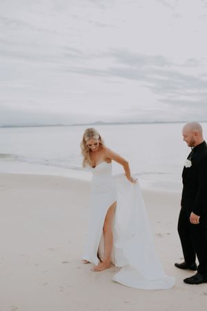 A playful moment as a bride lifts her wedding dress while walking on the beach.