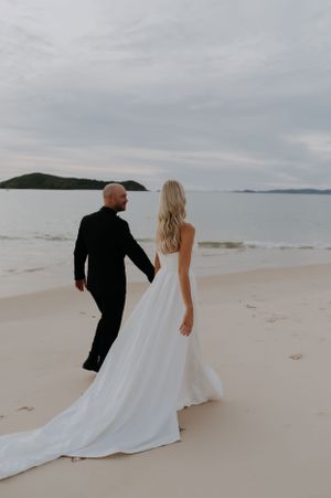 A newlywed couple walks along a pristine beach with mountains visible in the distance.
