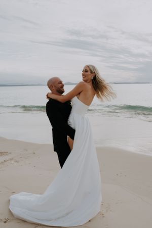 A romantic moment between a couple dancing on a beach at sunset with flowing white dress and black attire.