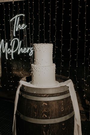 A white two-tier wedding cake displayed on a rustic barrel with a neon sign and fairy light backdrop.