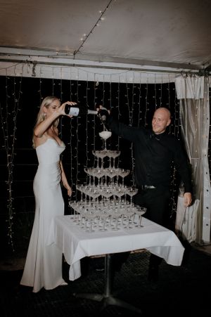 A couple pouring champagne into a tower of glasses at their wedding reception.