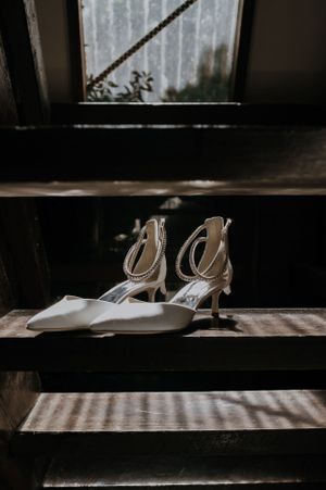 White bridal shoes with delicate straps sitting on wooden stairs by window.