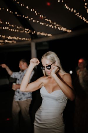 Party guest in white dress and sunglasses dancing under twinkling string lights at a nighttime celebration.