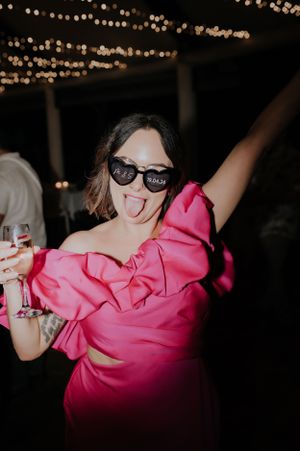 Woman in bright pink dress dances with drink in hand under string light decor.