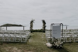 Outdoor wedding ceremony setup with white chairs and floral arch against cloudy sky.