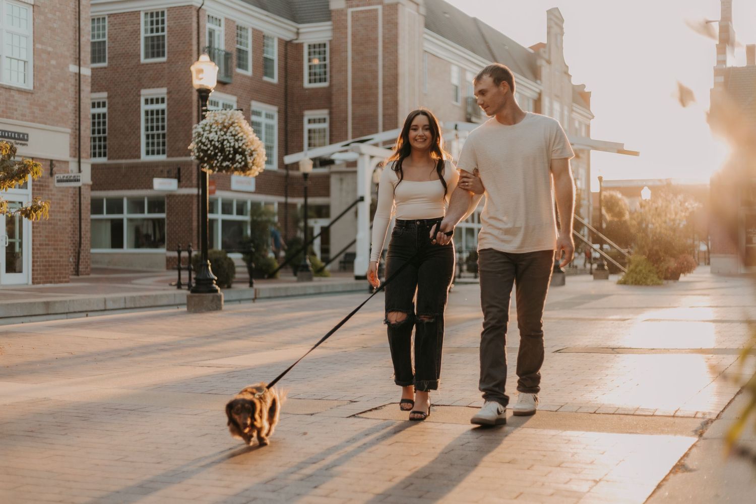 A couple walks their small dog down a city sidewalk during golden hour.