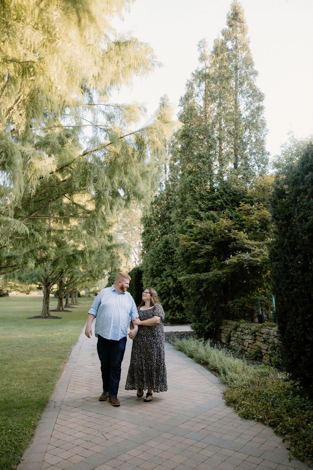 A couple walks together along a paved garden path surrounded by lush trees and greenery on a sunny afternoon.