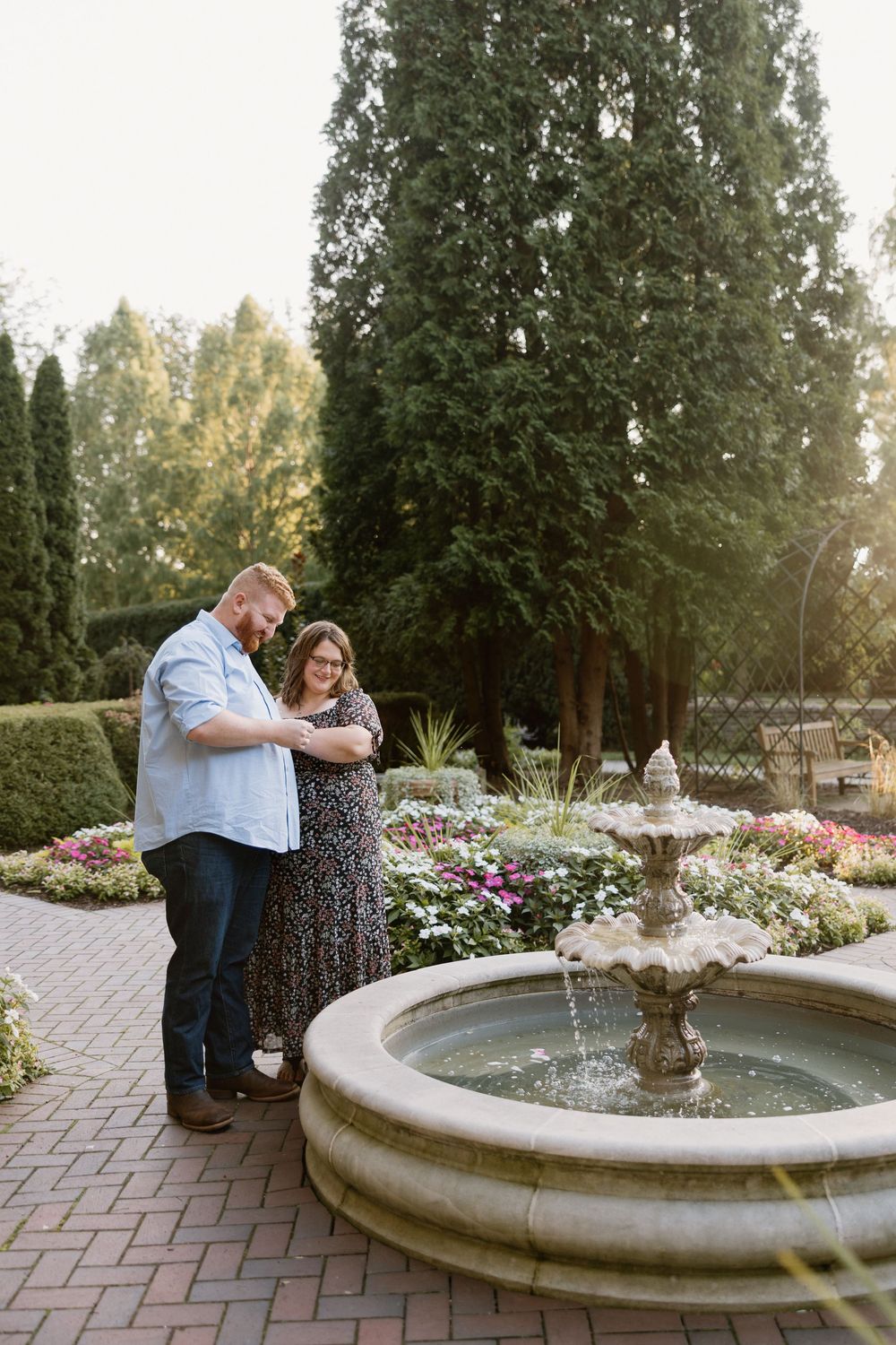 A couple shares an intimate moment beside a garden fountain surrounded by blooming flowers and trees.