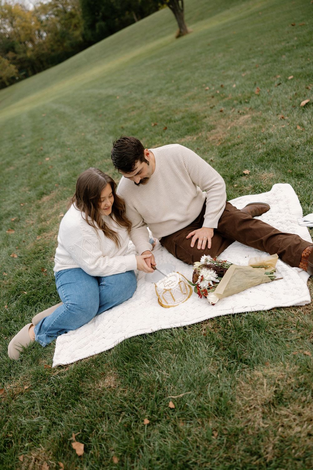A couple enjoys a cozy autumn picnic on a white blanket spread across a grassy hillside.