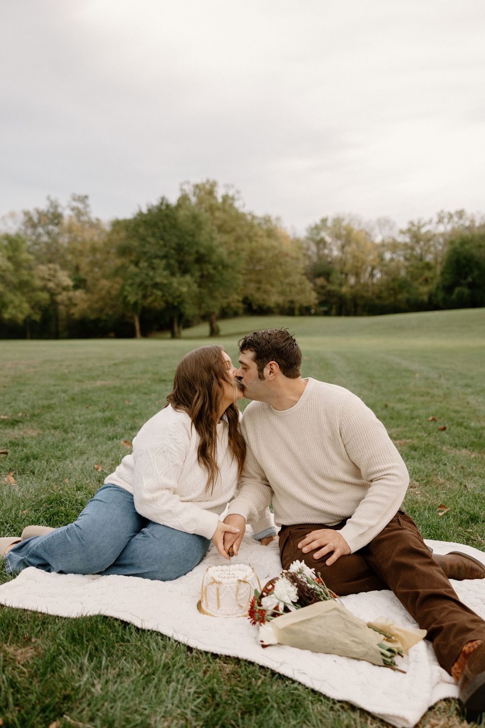 A couple shares a romantic moment while lying on a blanket in a grassy park setting during autumn.