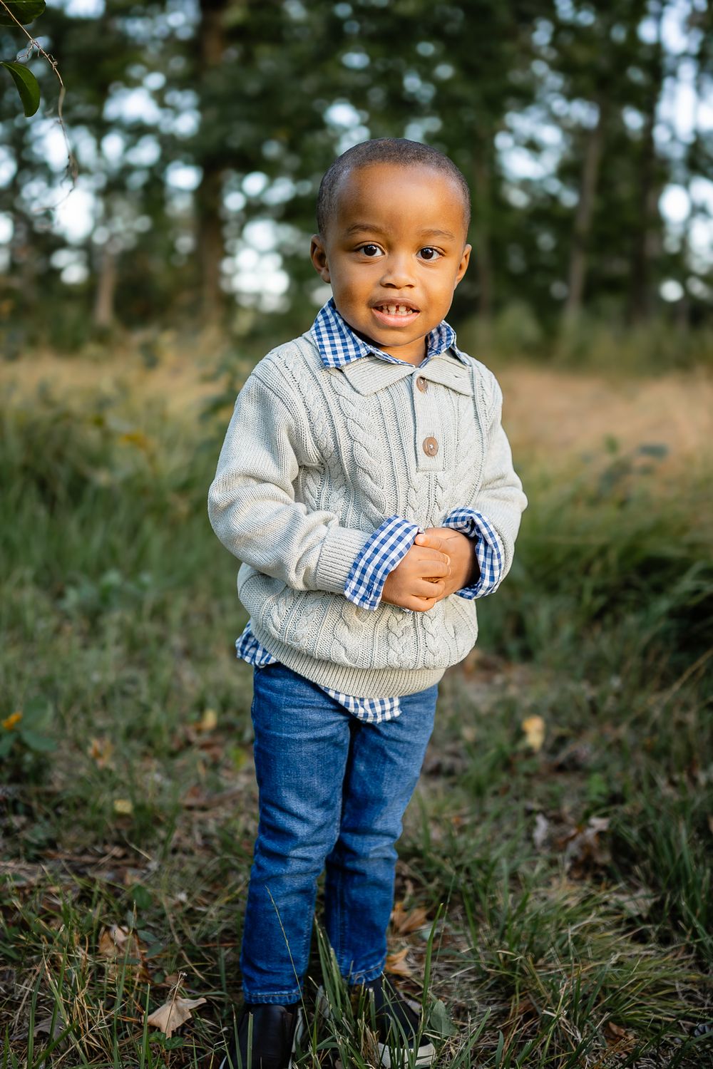 Young child in grey sweater and jeans stands in outdoor natural setting.