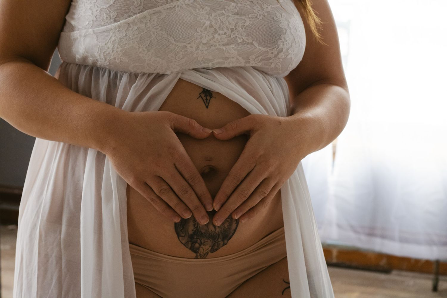 Hands form a heart shape over a pregnant belly in soft window light.