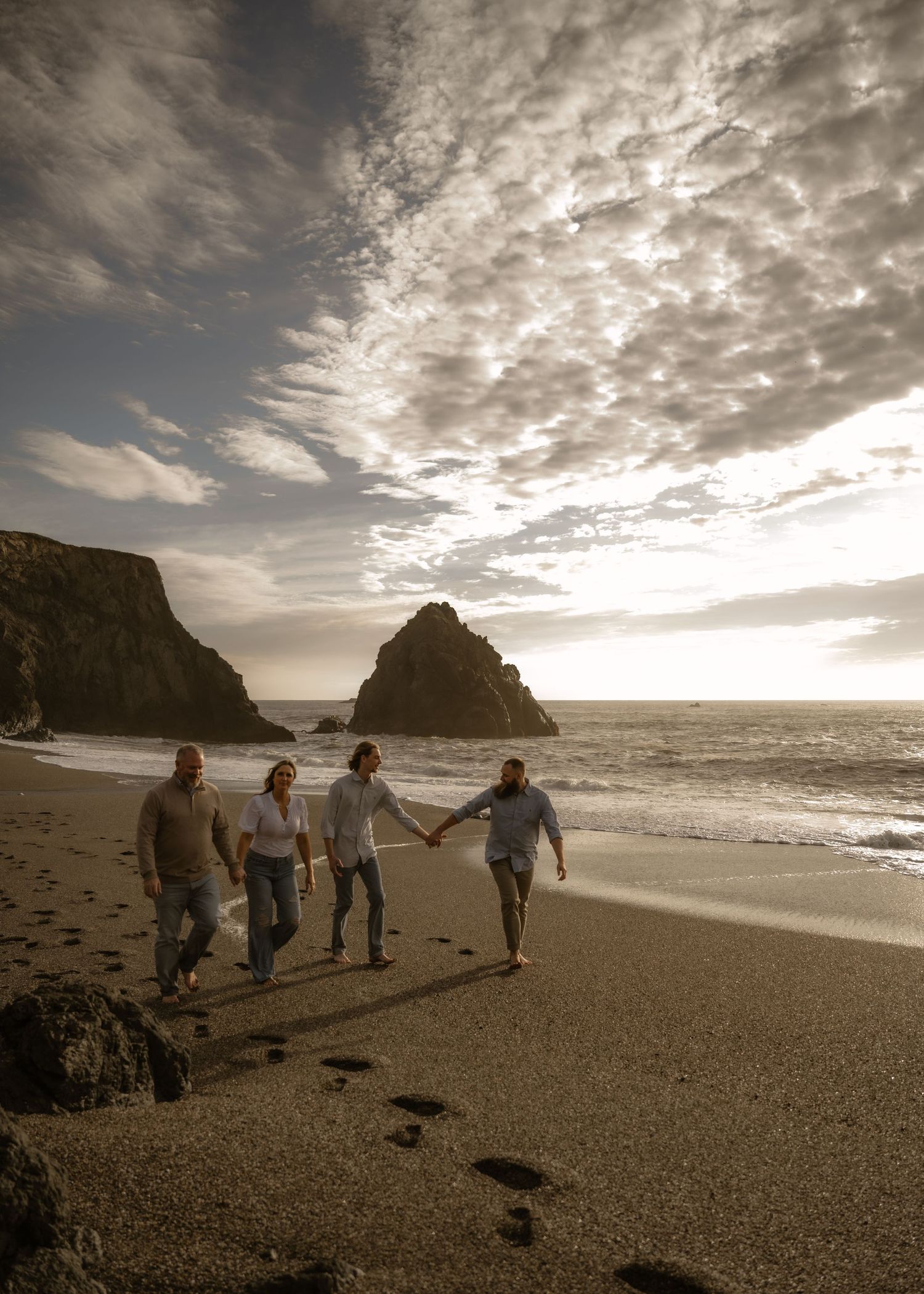 Four people stroll along a scenic beach with dramatic cliffs and cloudy skies at sunset.