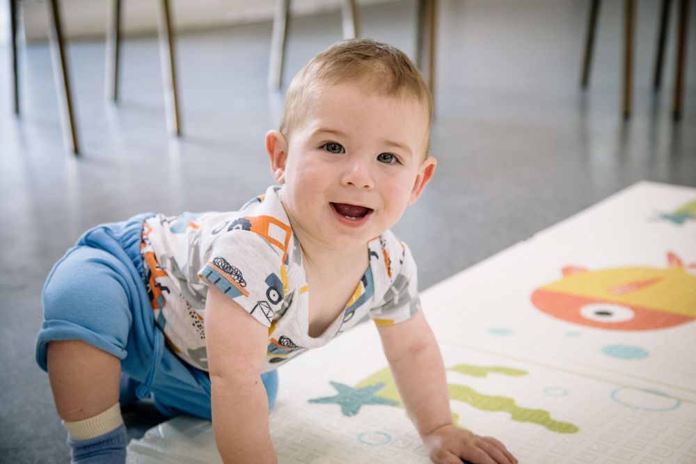 Baby in patterned shirt and blue shorts crawls on colorful playmat.