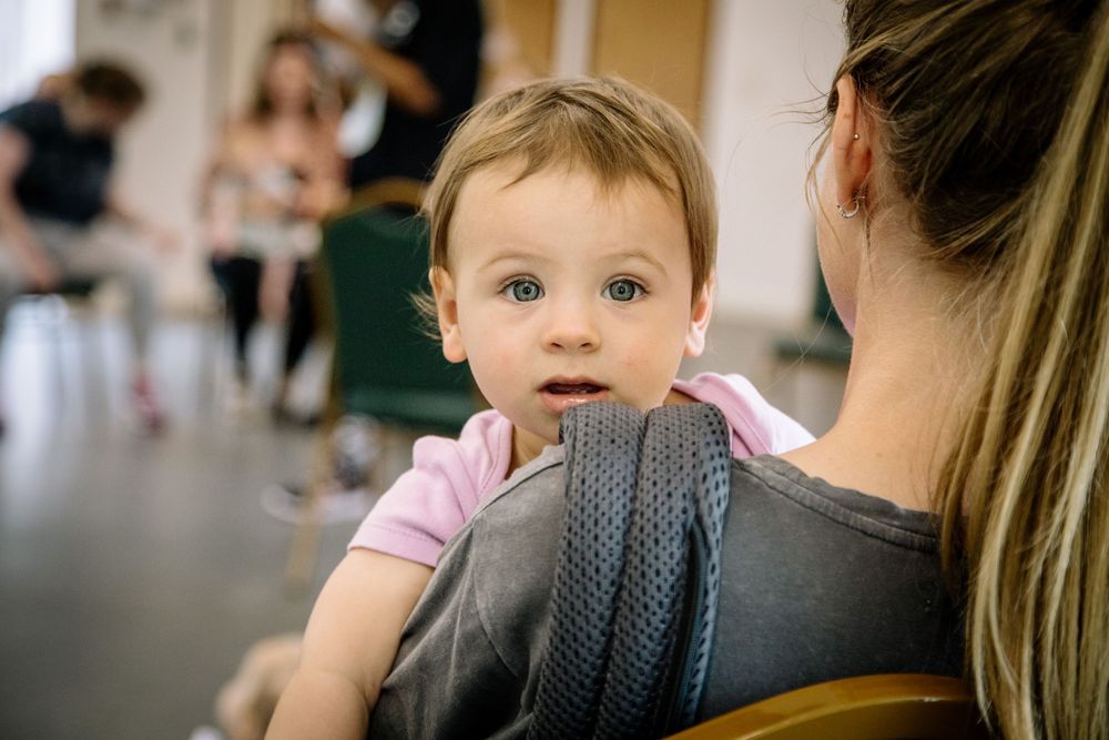 A baby in a pink shirt peers over a parent's shoulder while being carried in a dark baby carrier.