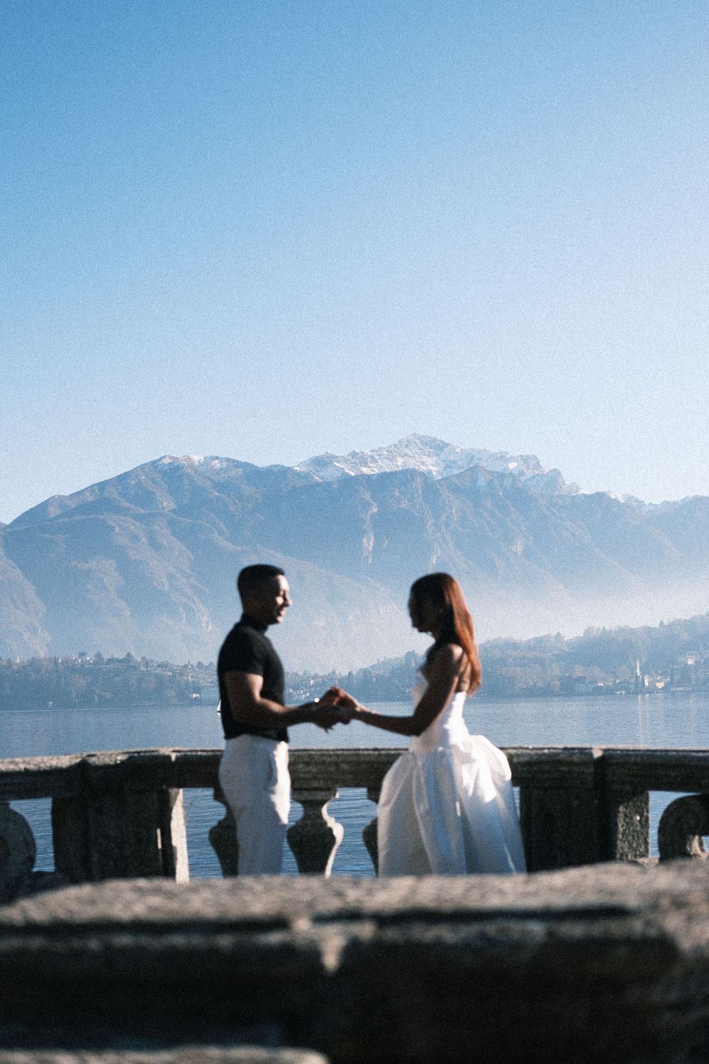 Two silhouettes stand together against a mountain backdrop at sunset near a stone wall overlooking a scenic landscape.