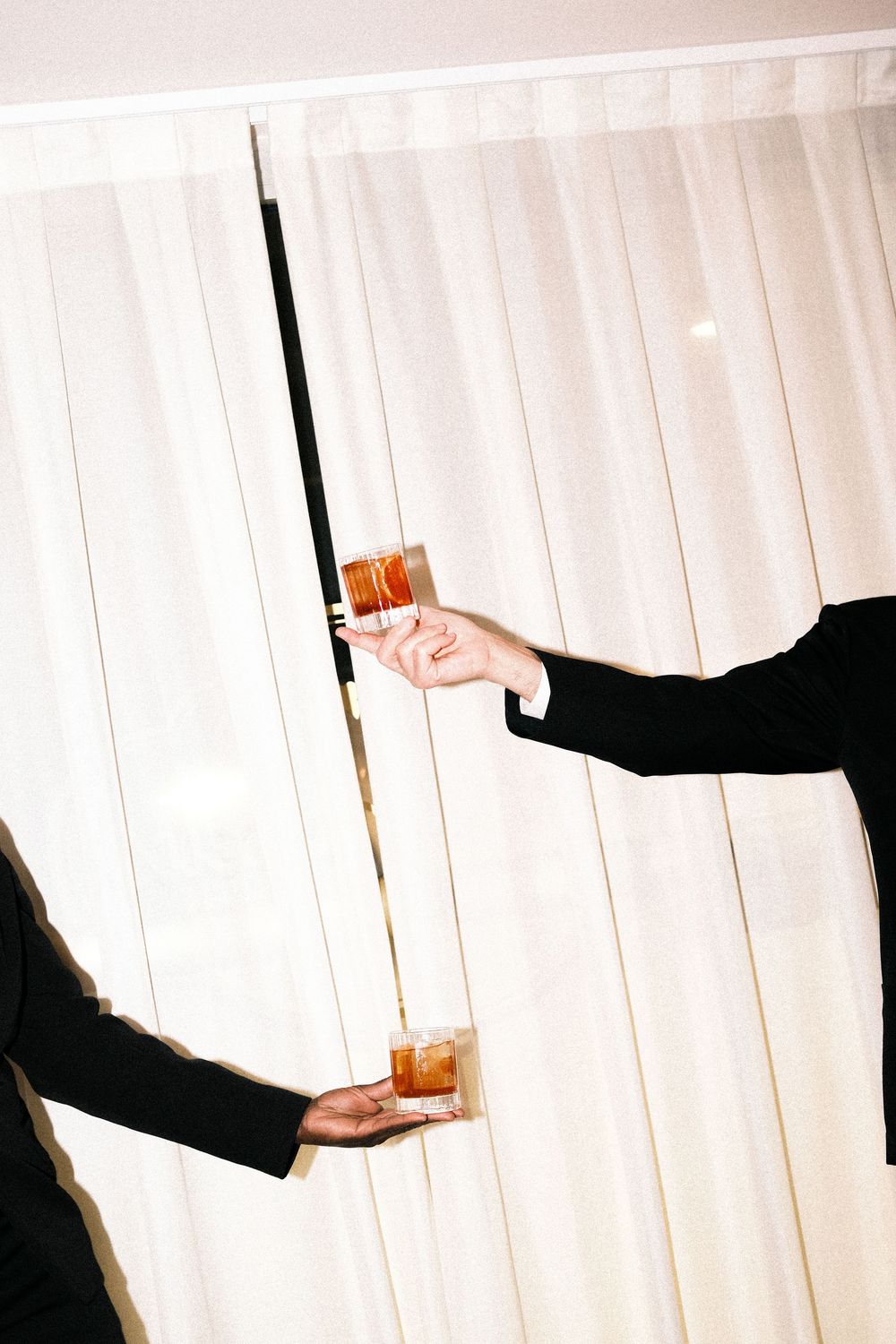 Two outstretched arms in dark suit sleeves hold glasses of amber cocktails against sheer white curtains in a bright, elegant room.