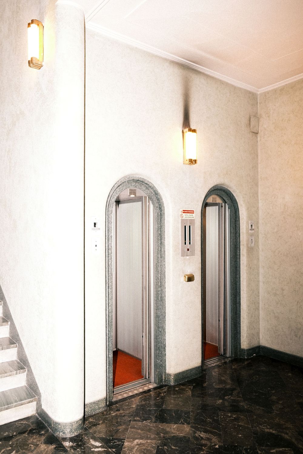 Two arched elevator doors with orange floors in a marble-trimmed lobby with wall sconces, dark polished stone flooring, and a staircase.