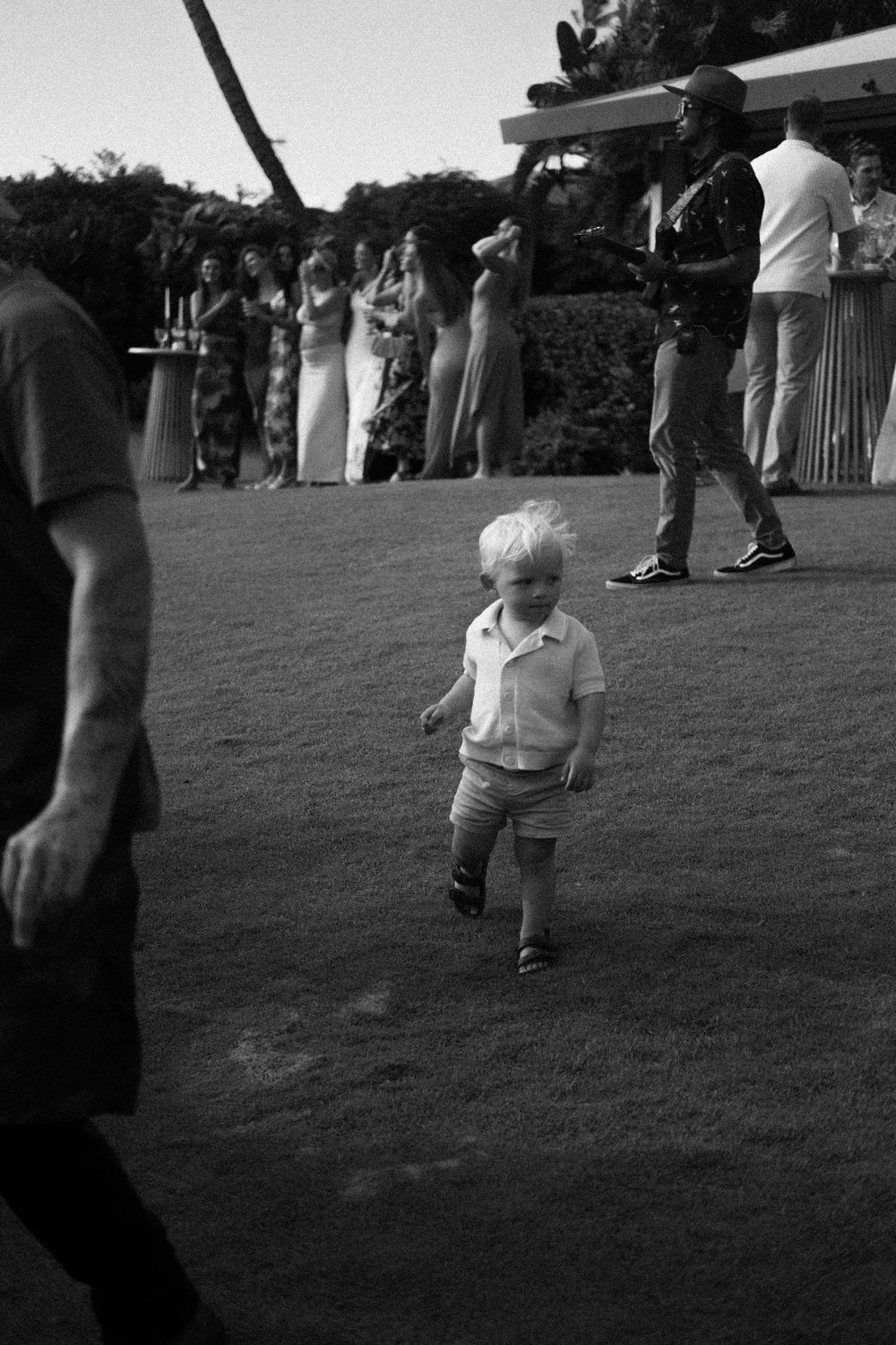 Black and white photo of a blond toddler walking across a lawn at an outdoor event, with guests gathered in the background.