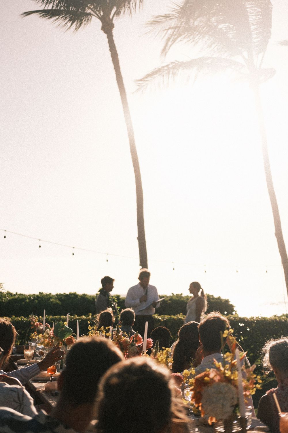 Guests seated at a flower-decorated outdoor wedding reception listen to a speech at golden hour beneath tall palm trees and string lights.