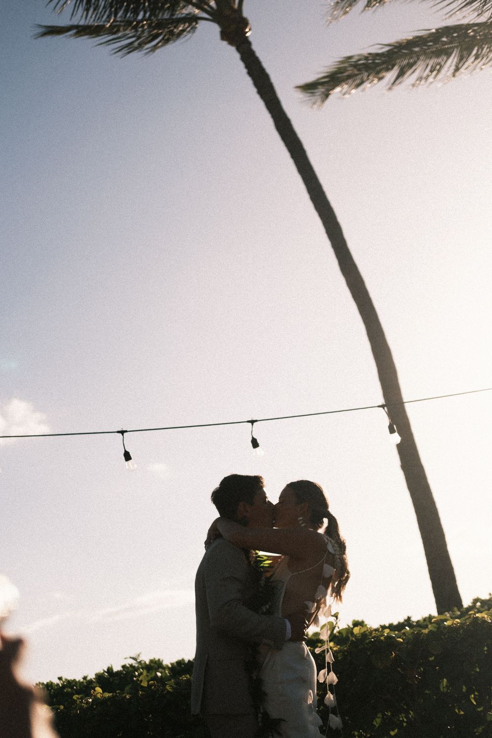 Couple sharing a romantic kiss at sunset under swaying palm trees with string lights at an outdoor tropical wedding.