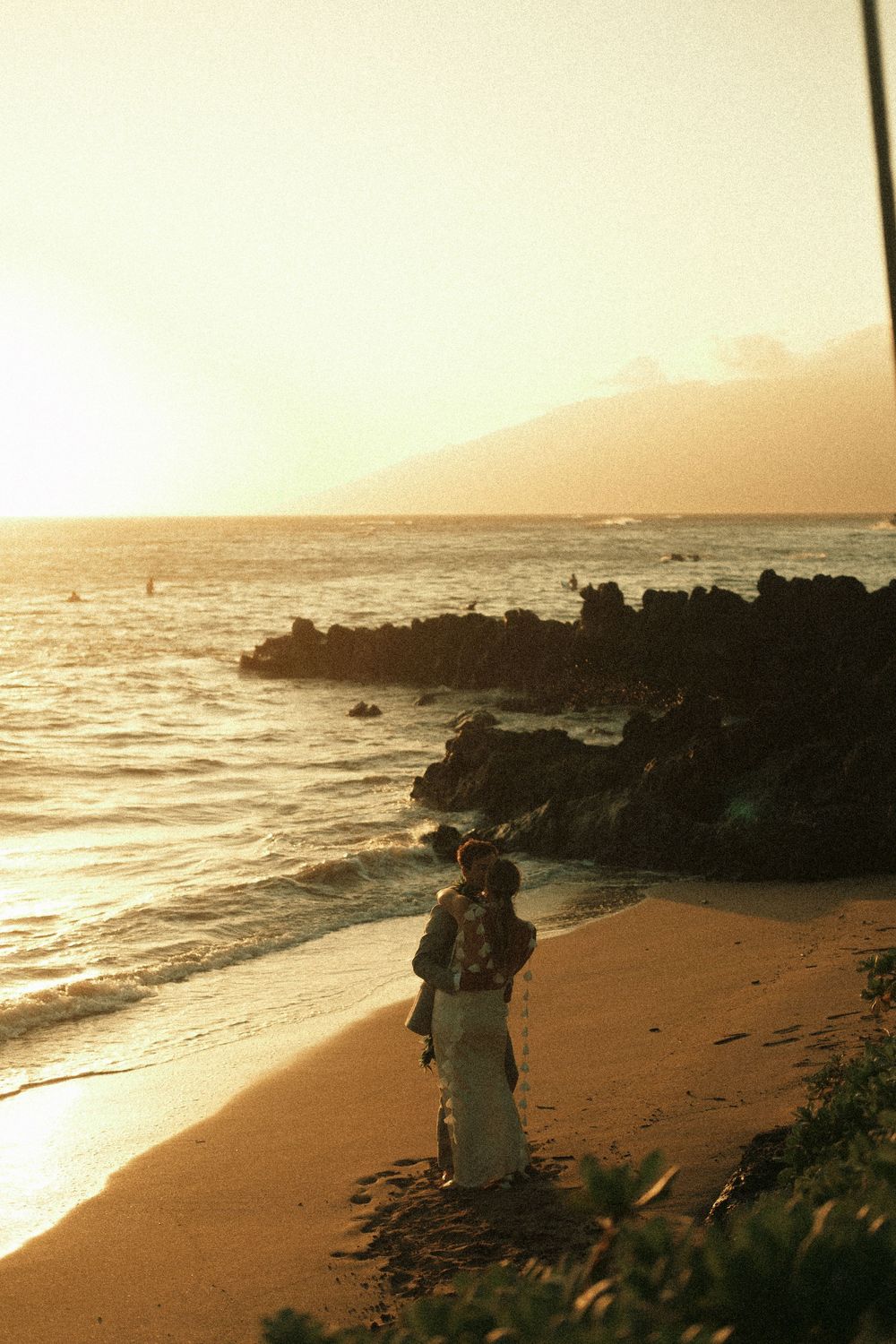 Couple embracing on a sandy beach at golden sunset with rocky shoreline and ocean waves in background.