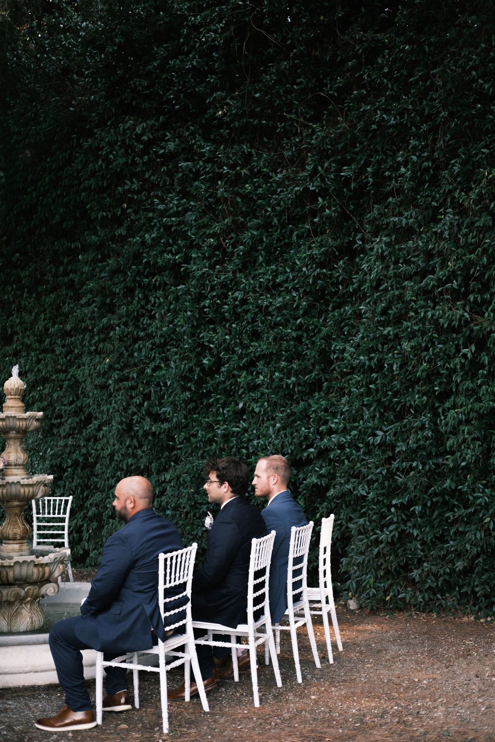 Three men in dark suits sit on white chairs facing a tall green ivy wall beside a tiered stone fountain at an outdoor wedding ceremony.