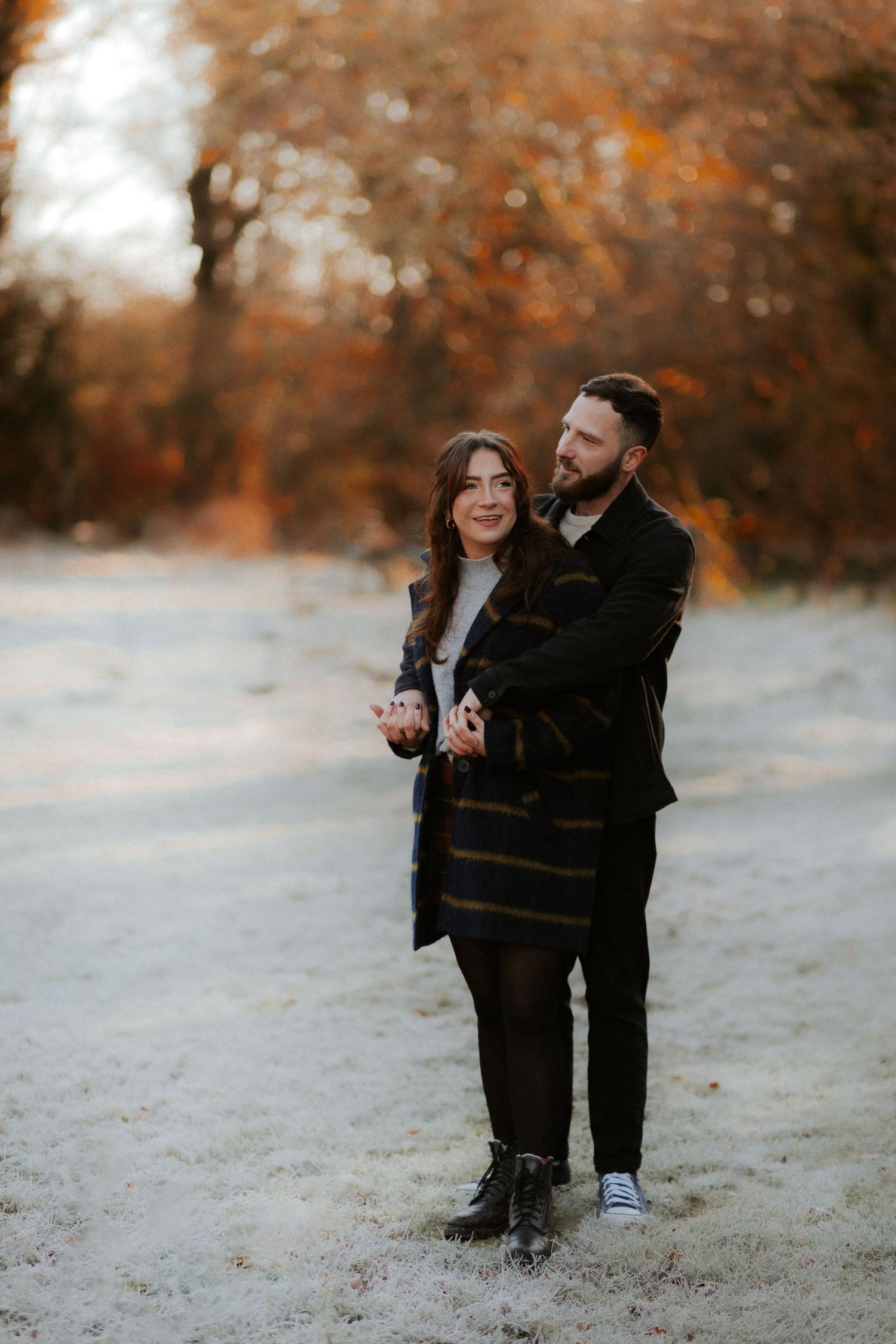 A romantic winter engagement photo of a couple embracing in a snowy landscape with autumn trees in the background.