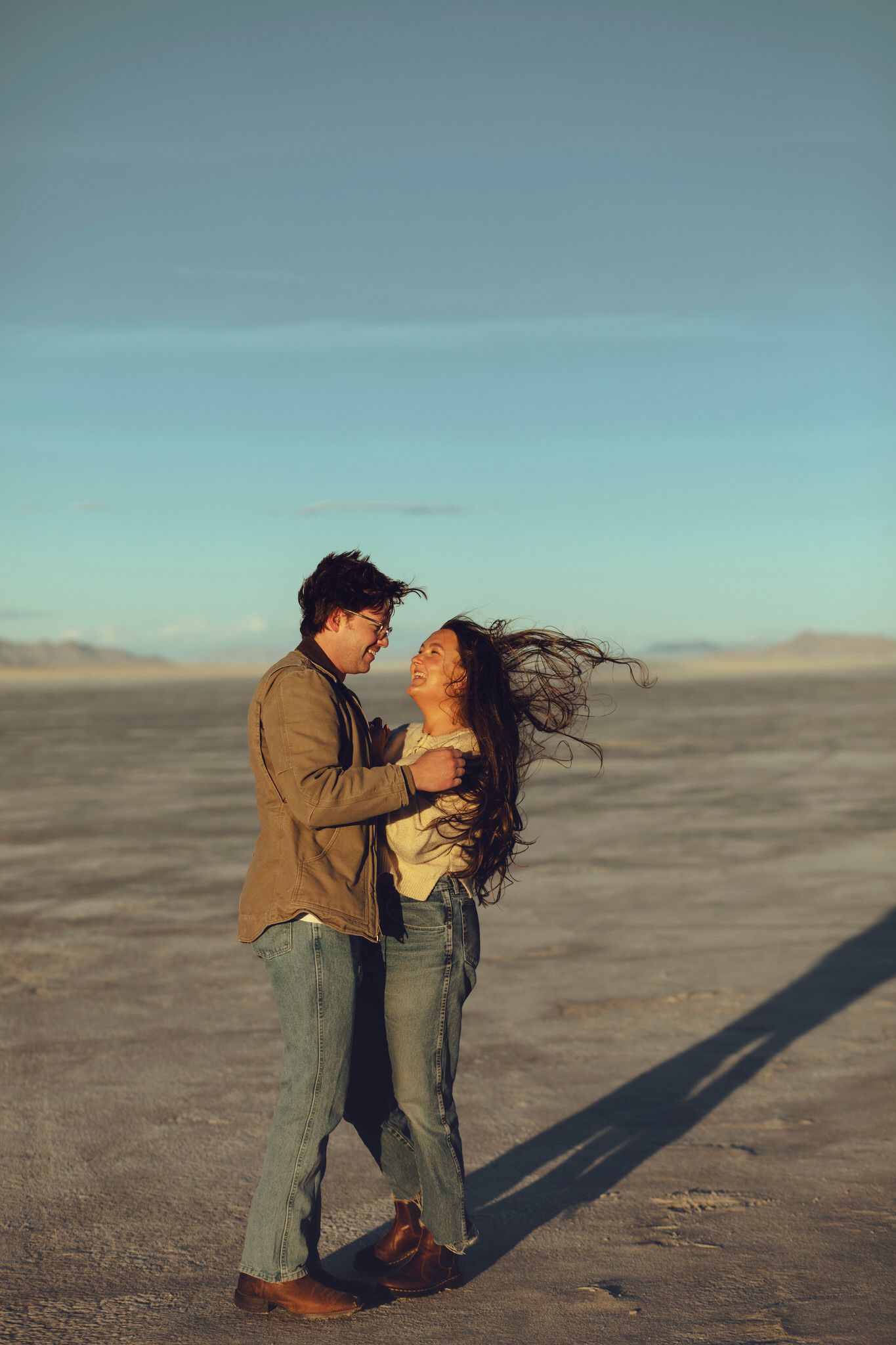 Couple standing and smiling at each other on the salt flats with their hair blowing in the wind.