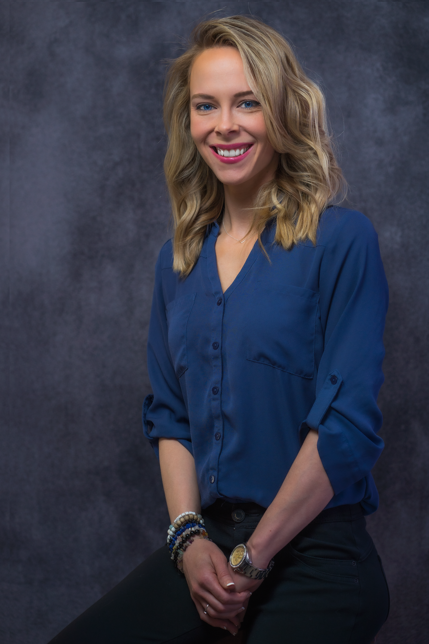 Professional portrait of someone in a navy button-up blouse and dark pants against a gray textured backdrop.