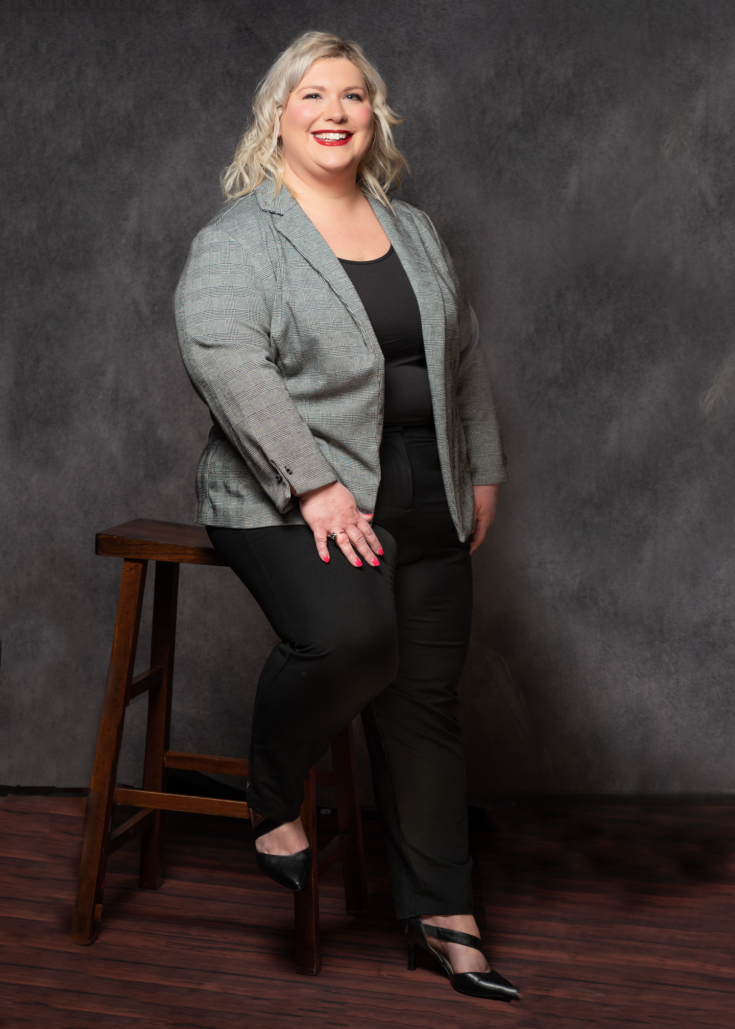 Professional studio portrait of a blonde woman in business attire leaning against a wooden stool against a dark backdrop.