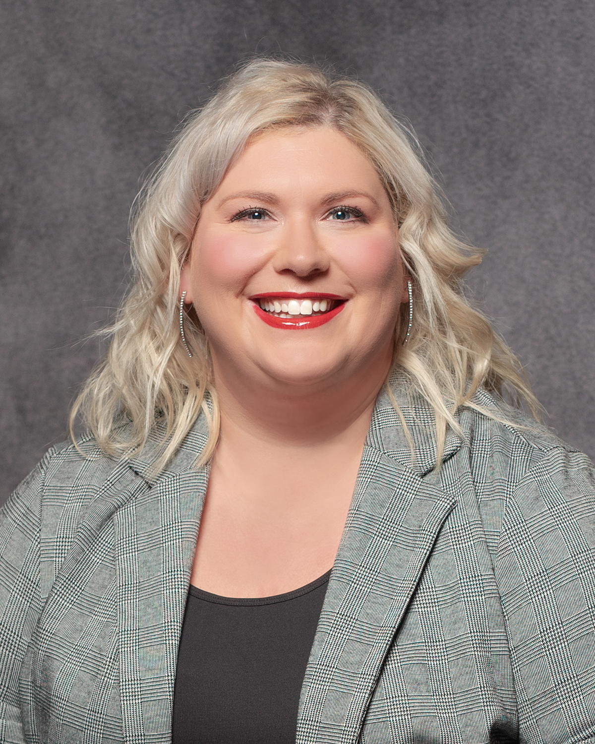 Professional headshot showing a person with blonde wavy hair wearing a gray blazer and black top, smiling warmly against gray background.