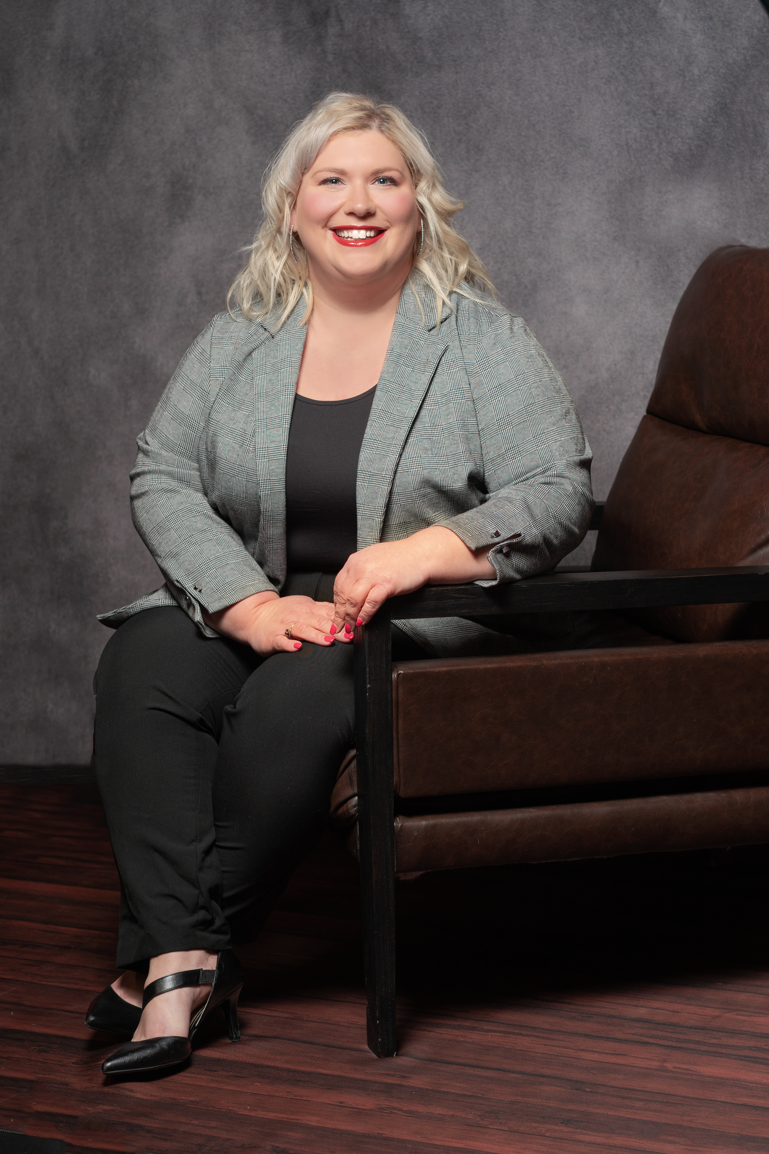 Professional headshot of someone in a gray blazer and black outfit seated on a brown couch against a gray backdrop.