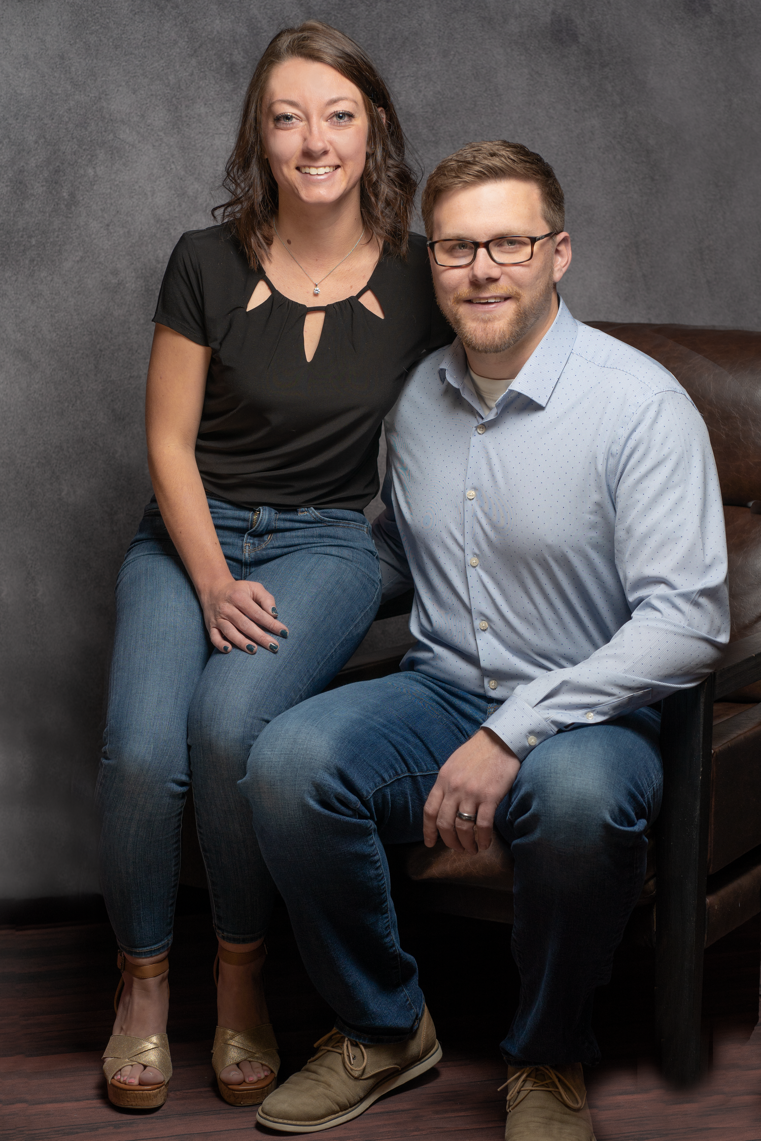 A couple sits together on a brown leather chair wearing casual attire against a gray studio backdrop.
