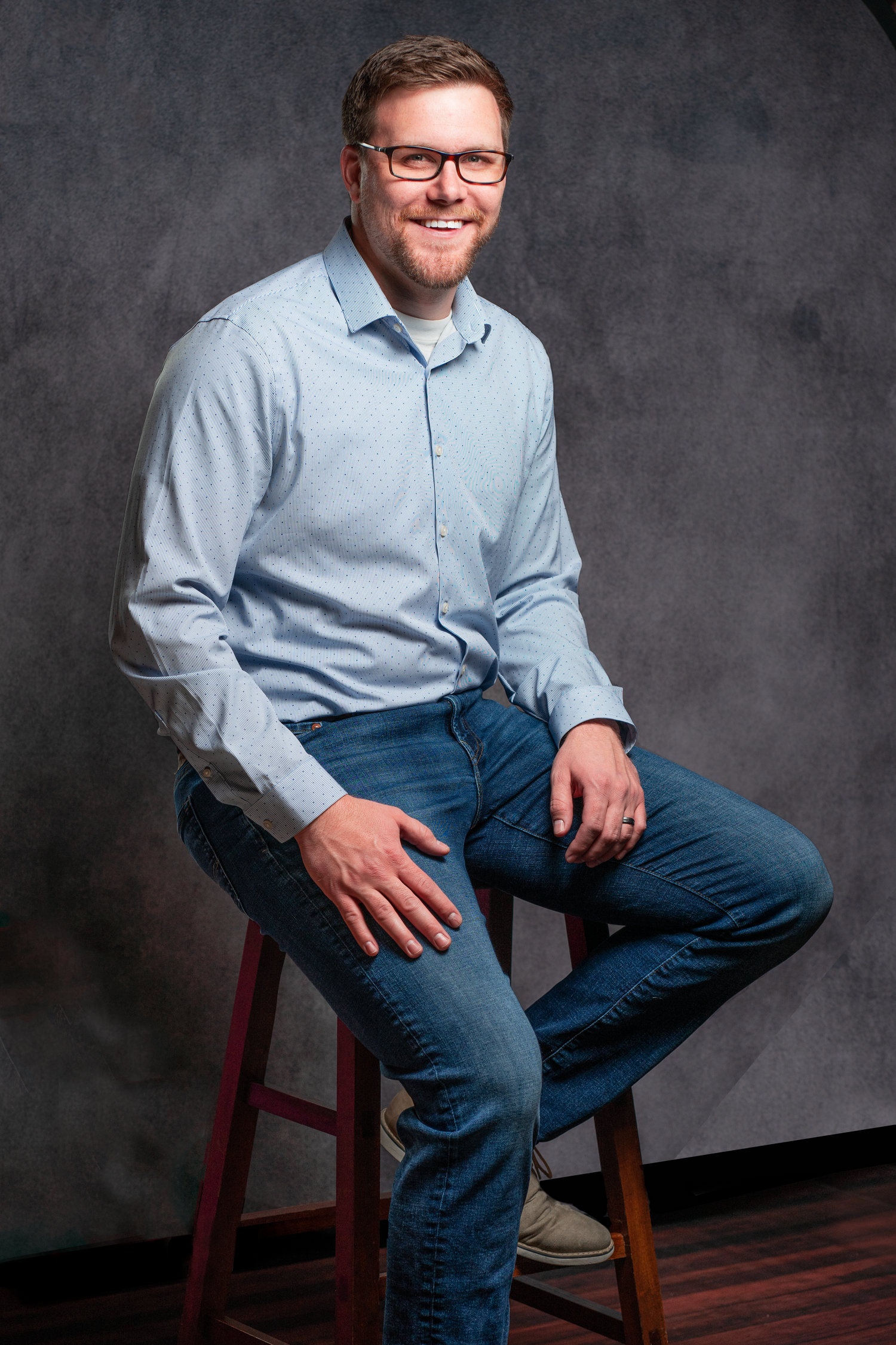 Portrait of a person in a light blue button-down shirt and jeans sitting casually on a wooden stool against a gray backdrop.