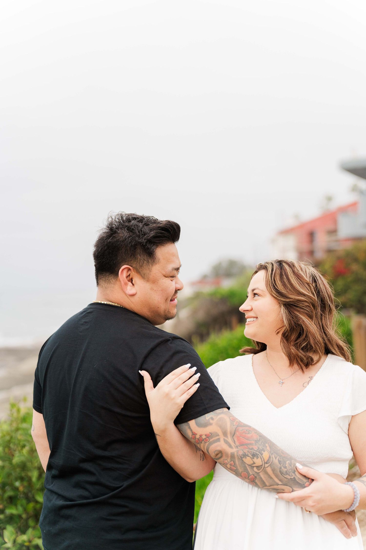 A couple in black and white outfits share a loving moment outdoors against a cloudy sky.
