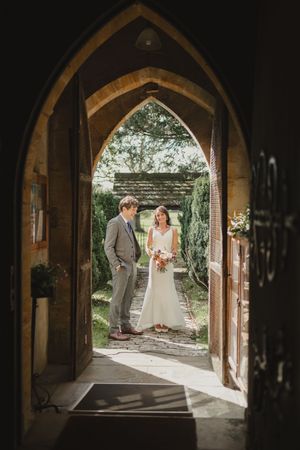 Wedding couple framed in gothic church archway with garden view beyond.