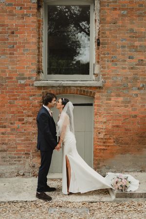 Couple in wedding attire embraces against a rustic brick wall backdrop.