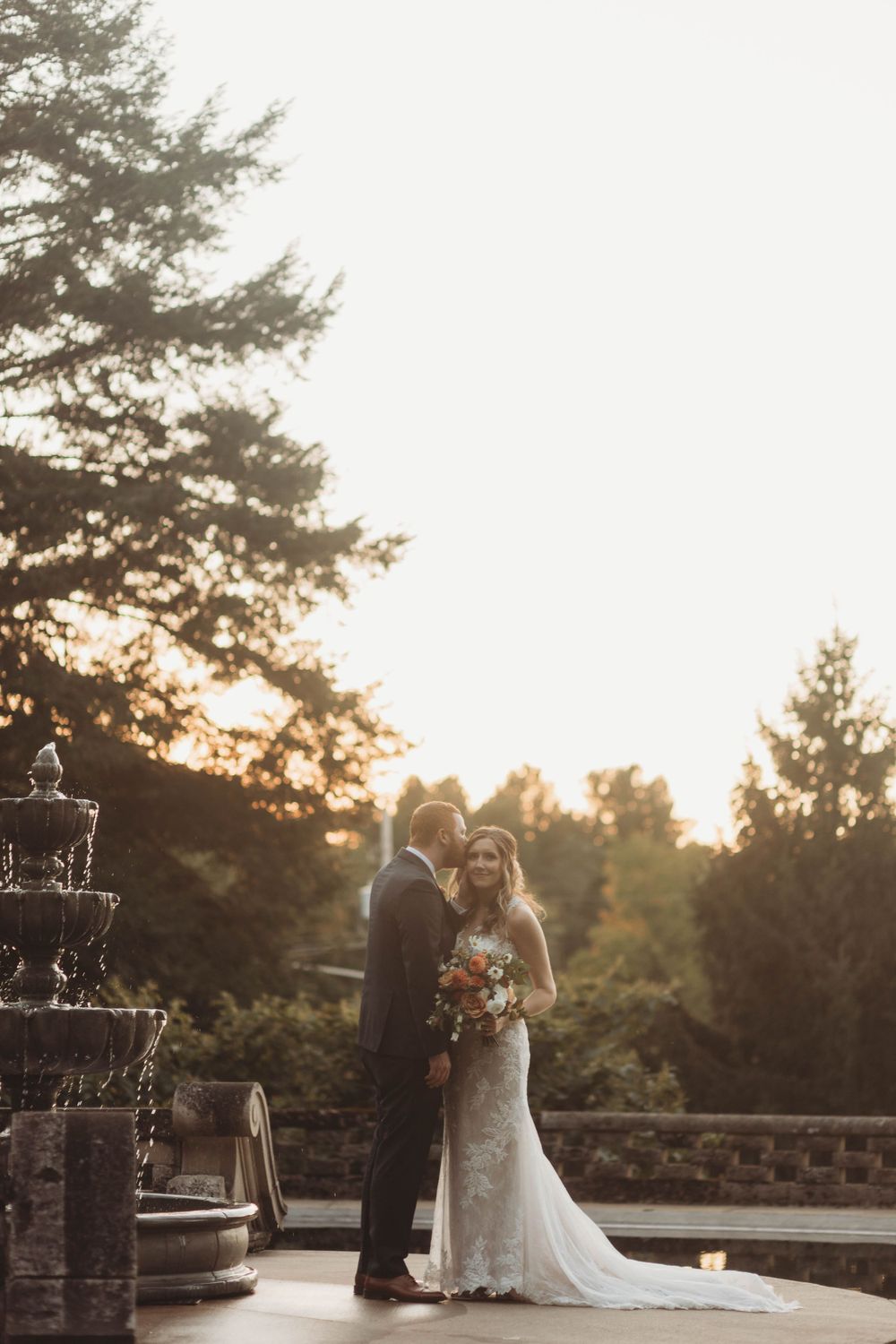 Wedding couple shares romantic moment at sunset with pine trees and stone architecture.