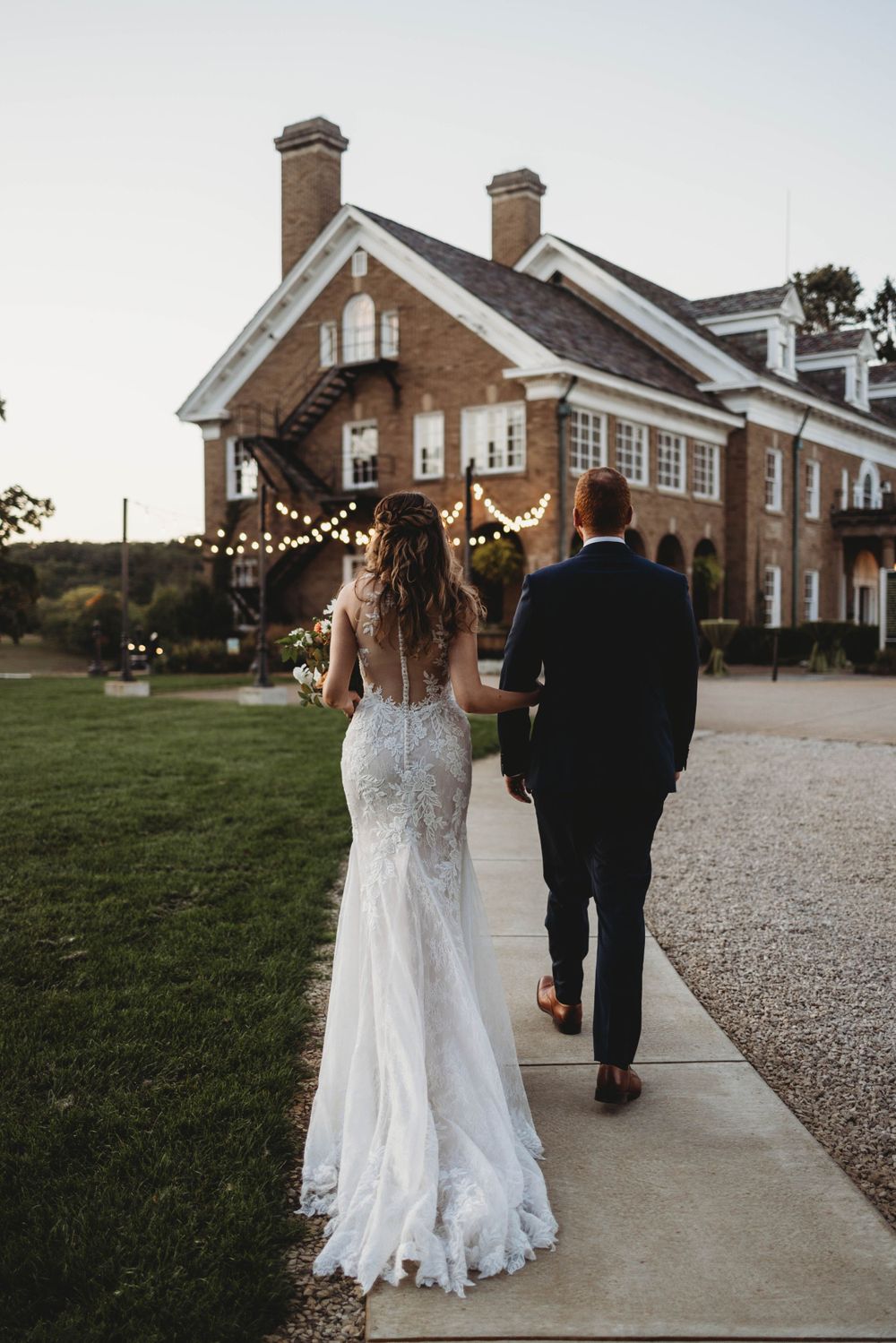 Wedding couple walks along gravel path towards brick building with string lights at dusk.